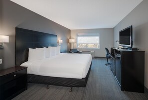 A modern hotel room with a large bed, a desk with a computer, a television, and a window with blinds.