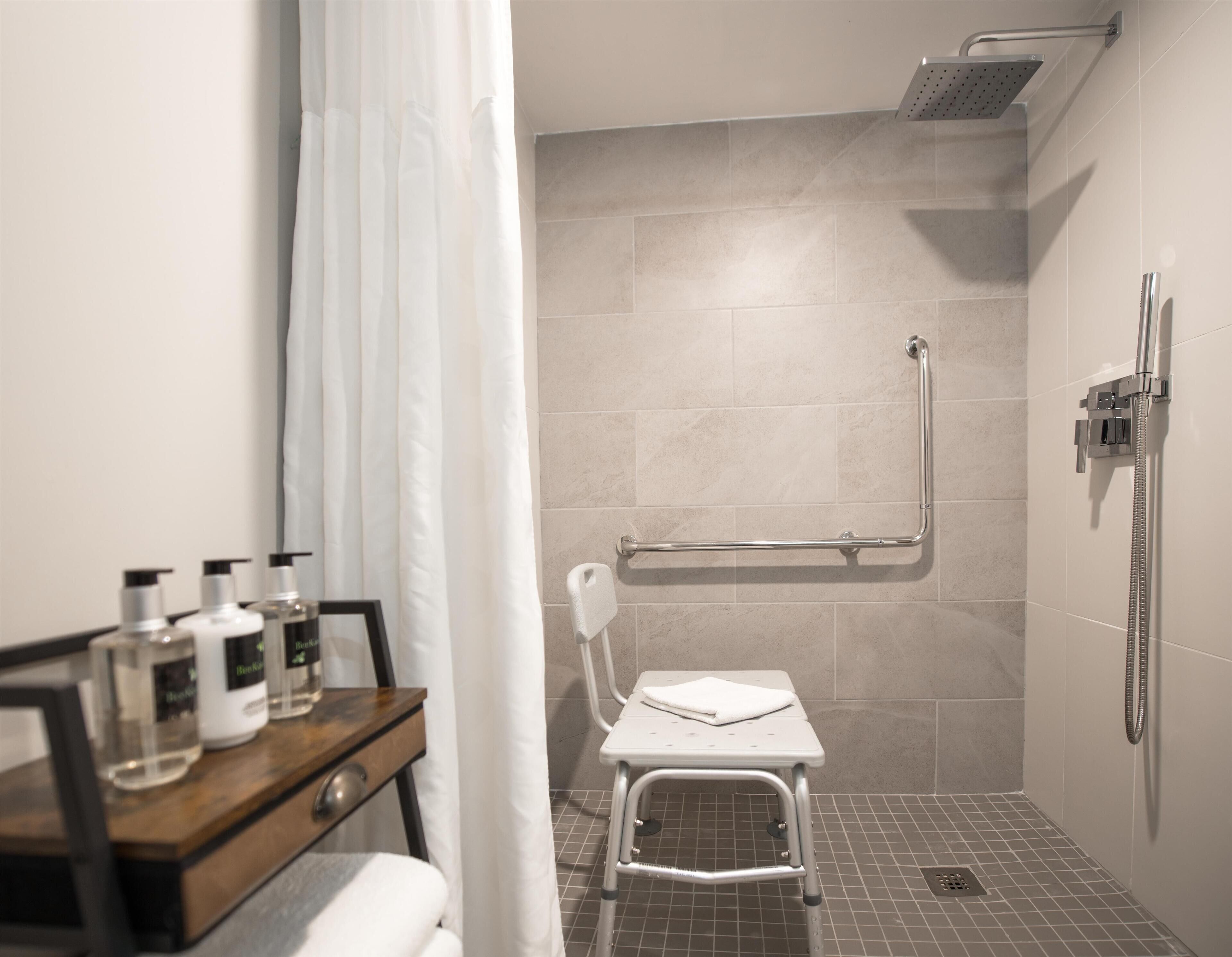 Un baño moderno con ducha, barras de apoyo, una silla blanca y artículos de aseo en un mostrador de madera.