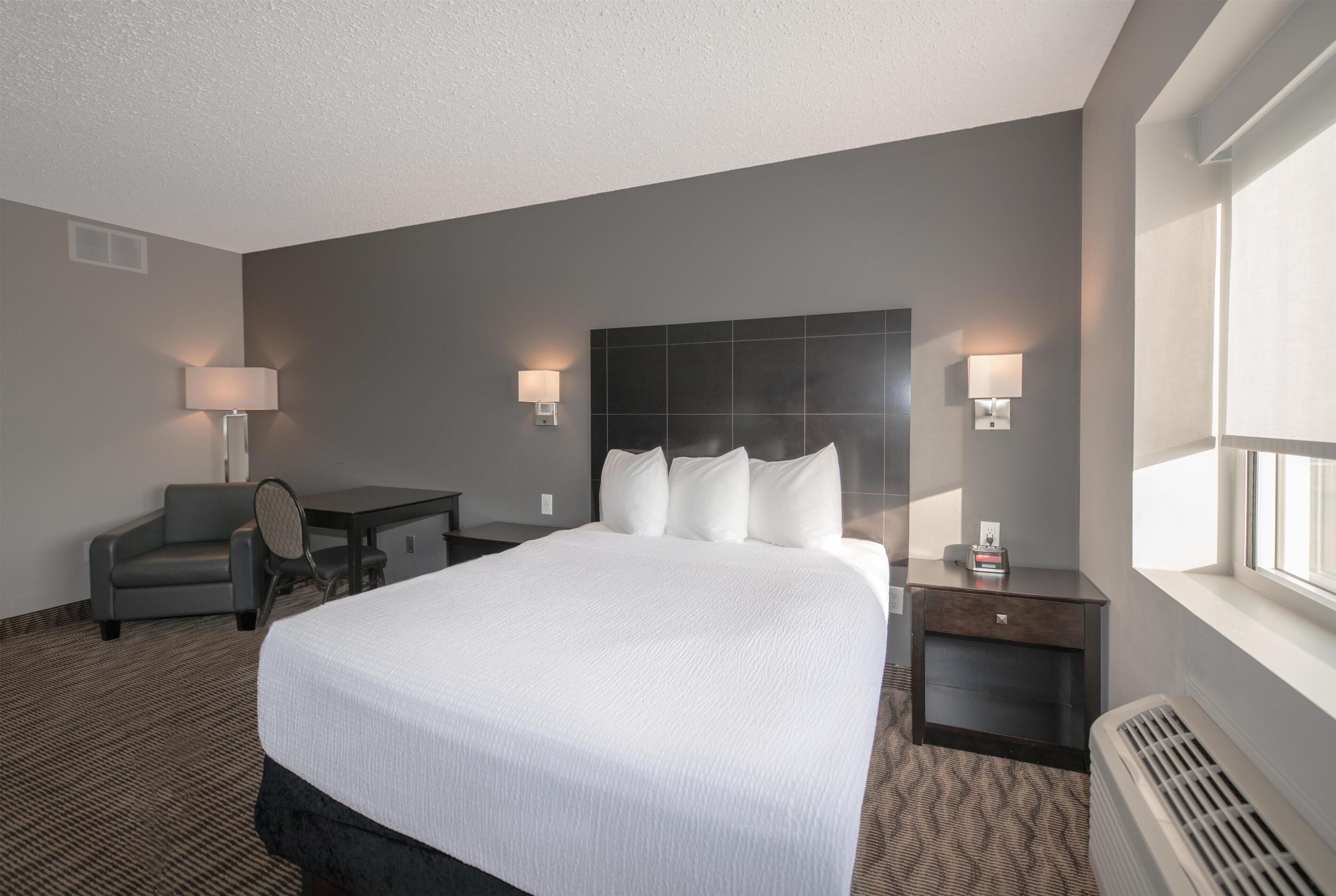 Une chambre d’hôtel avec un grand lit, deux tables de chevet avec des lampes, un bureau avec une chaise et une fenêtre avec des stores.