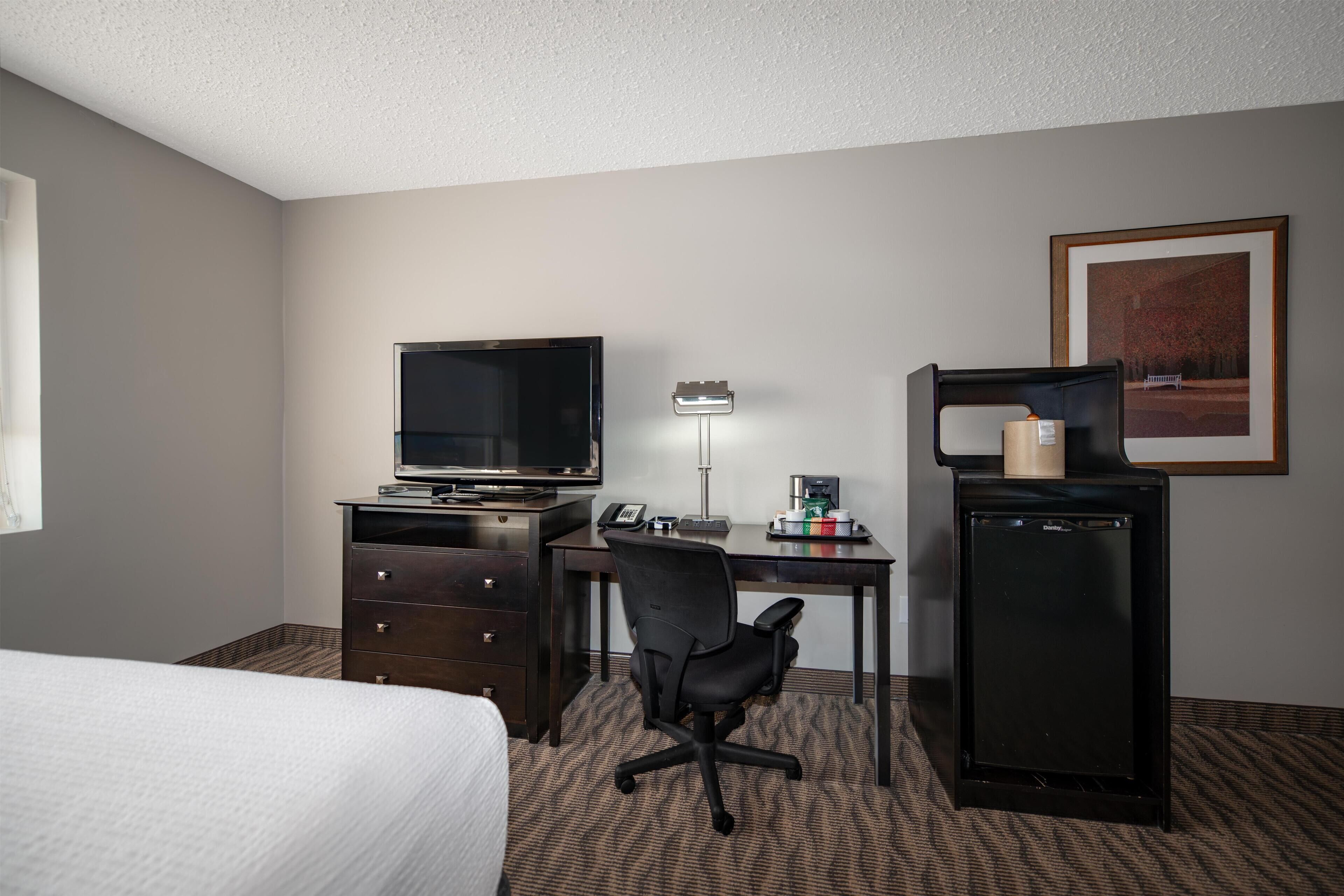 Quarto de hotel com cama, escrivaninha com computador, televisão, frigorífico e luminária.