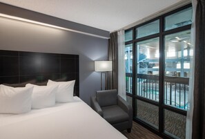 Habitación de hotel con una cama grande, una silla, una lámpara y vistas a un espacio exterior a través de amplios ventanales.