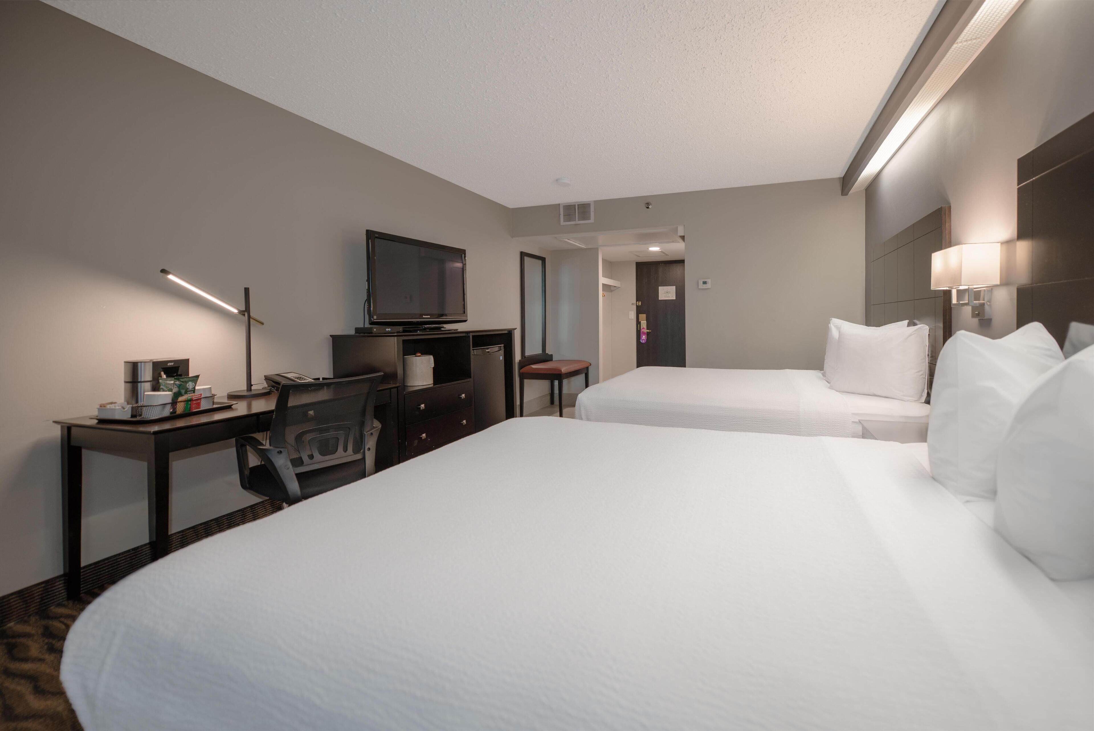 Quarto de hotel com duas camas, uma escrivaninha com computador, televisão e luminária.