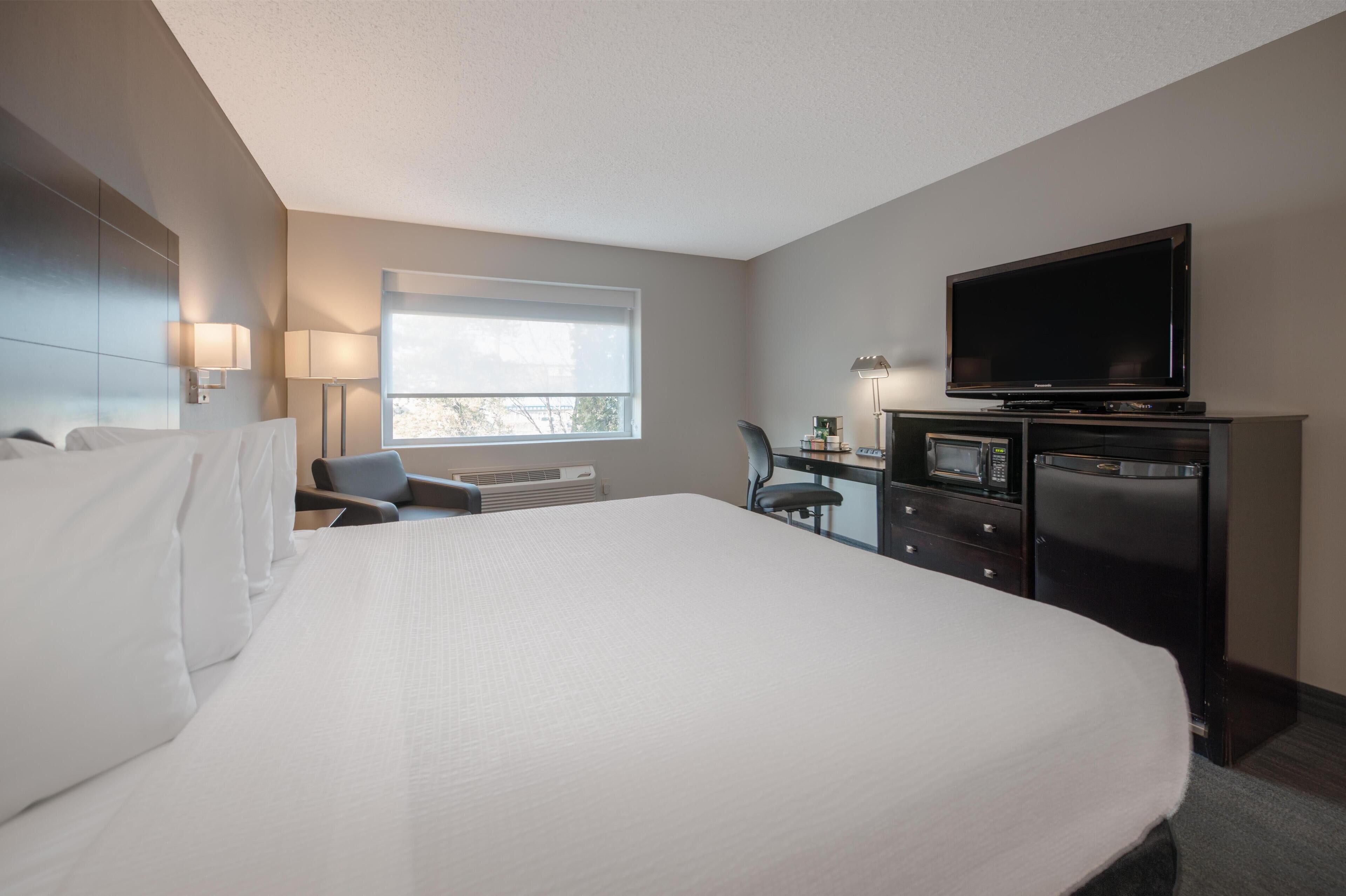 A hotel room with a large bed, a desk with a television, a chair, and a window with blinds.