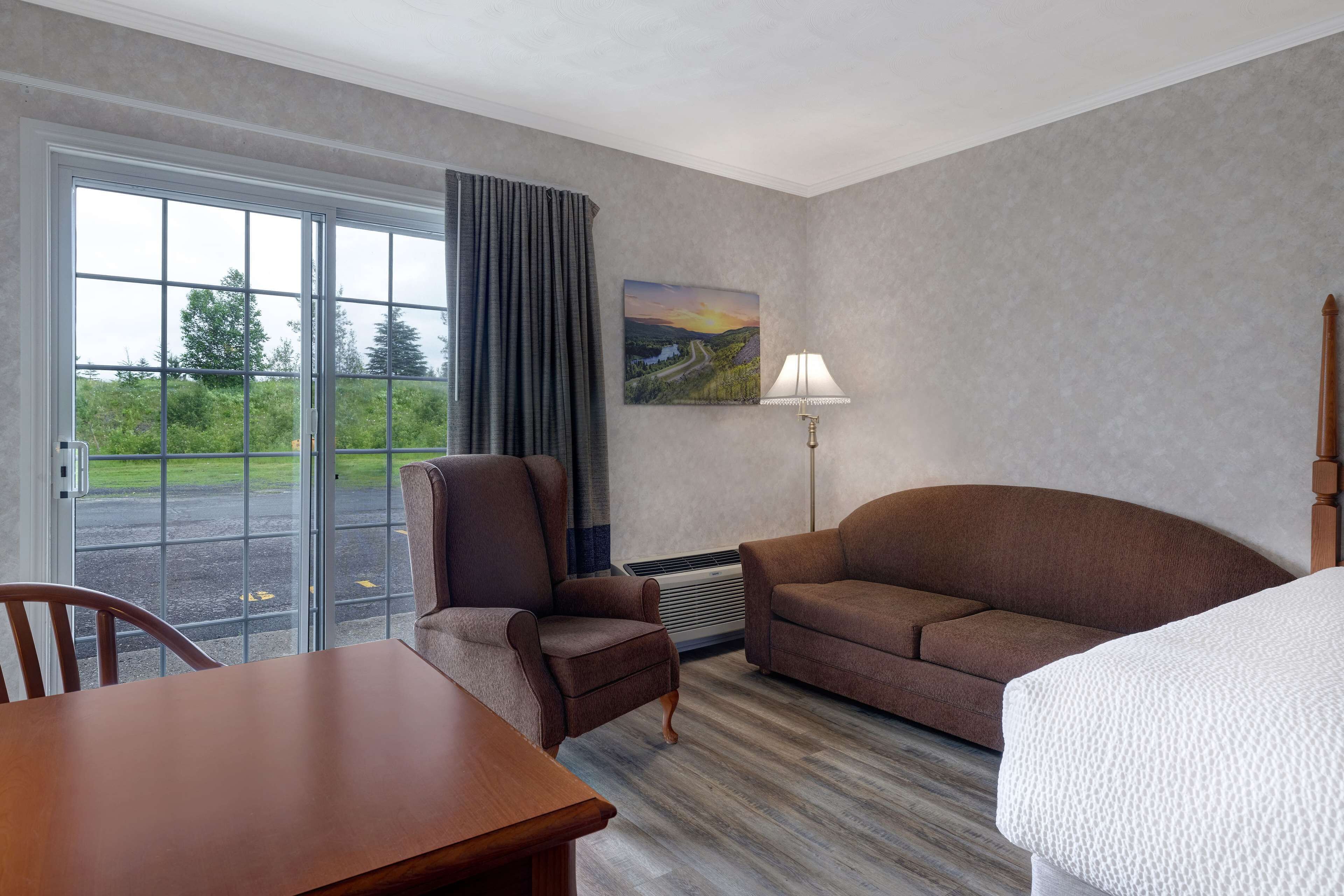 Habitación de hotel con un amplio ventanal corredera, un sillón marrón, un sofá marrón, una mesa de madera y una cama blanca.