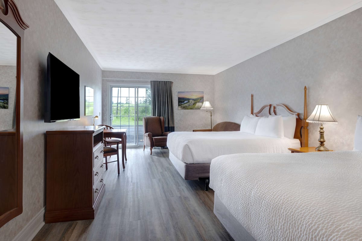 Habitación de hotel con una cama grande, una cama más pequeña, un escritorio, una silla, un televisor y una ventana con vista a follaje.