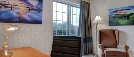A hotel room with a wooden table, a black office chair, a brown armchair, a floor lamp, and two paintings on the walls.