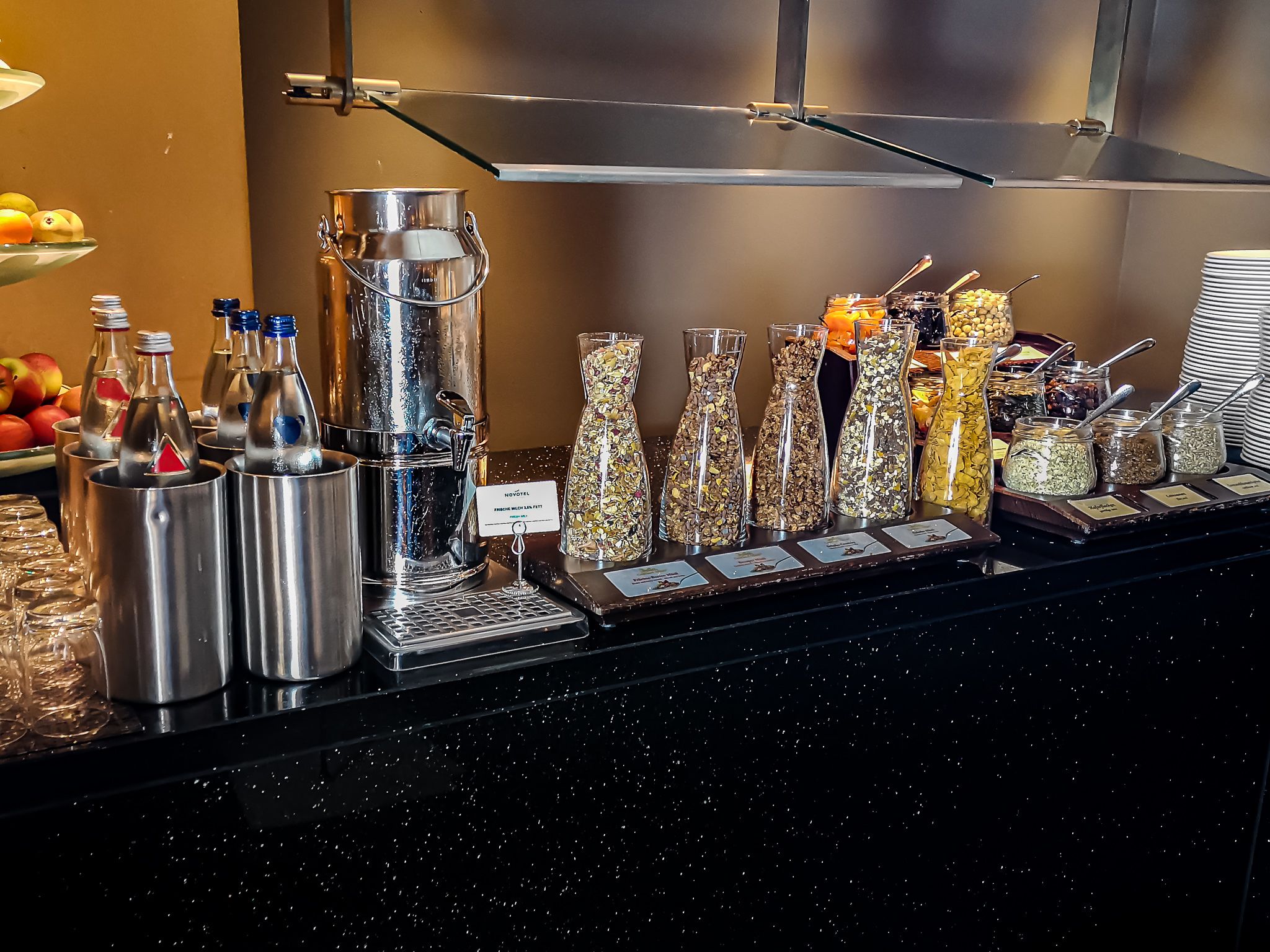 Un buffet composé de différents récipients contenant des aliments, notamment un grand distributeur en métal, des bocaux en verre contenant divers produits alimentaires et des plats de service étiquetés.