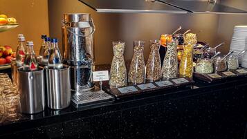 Un buffet composé de différents récipients contenant des aliments, notamment un grand distributeur en métal, des bocaux en verre contenant divers produits alimentaires et des plats de service étiquetés.