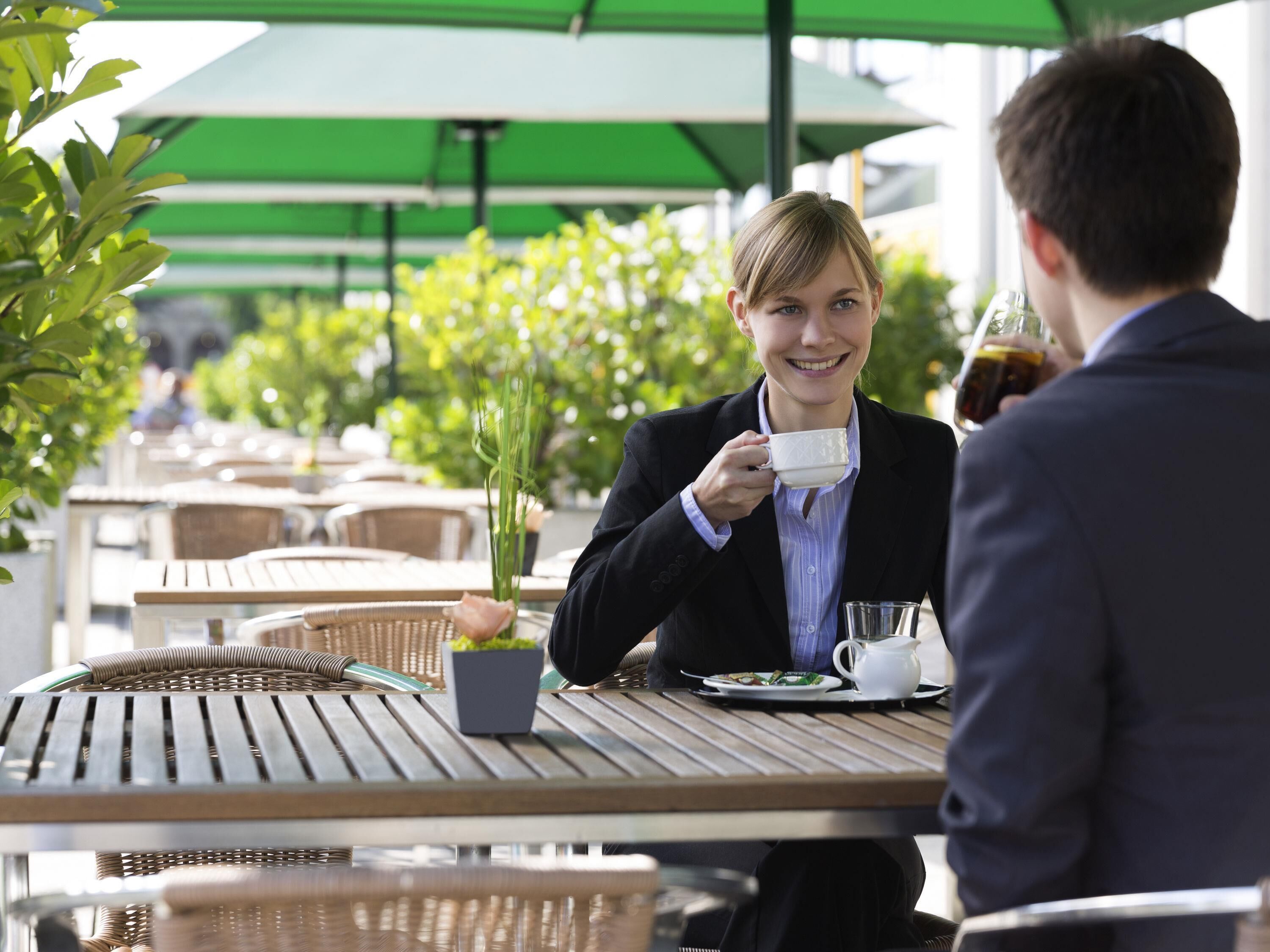 An outdoor café setting with tables and chairs, a person holding a cup, and another person seated across.