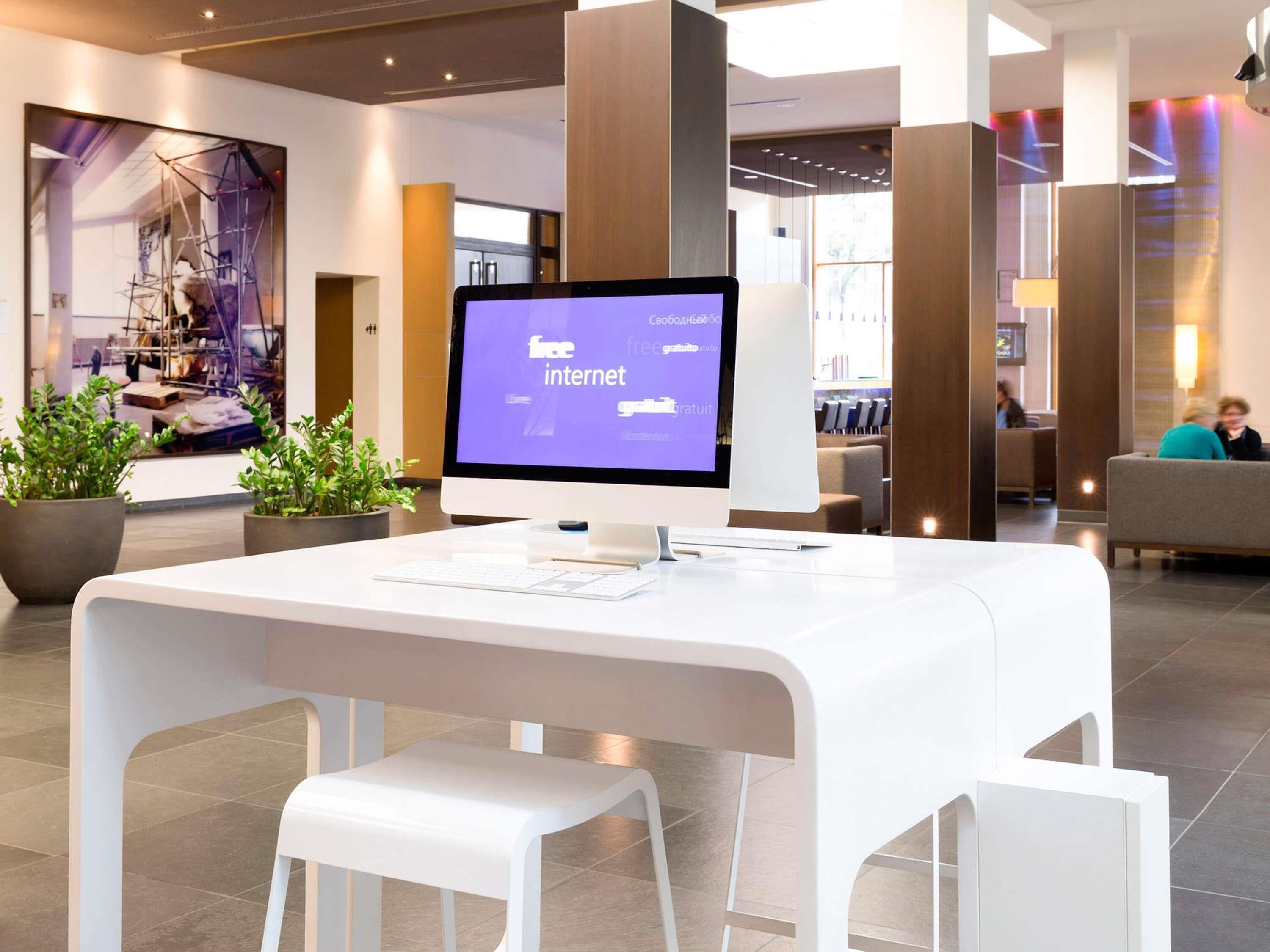 A modern hotel lobby with a white reception desk, a large monitor displaying a website, potted plants, and a seating area in the background.