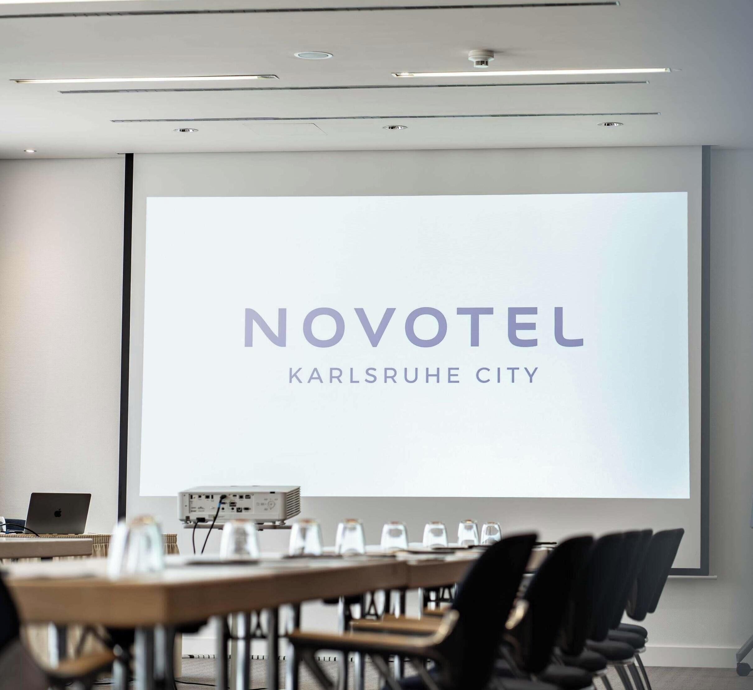 A conference room with a projection screen displaying 'NOVOTEL KARLSRUHE CITY' and a laptop on a table.