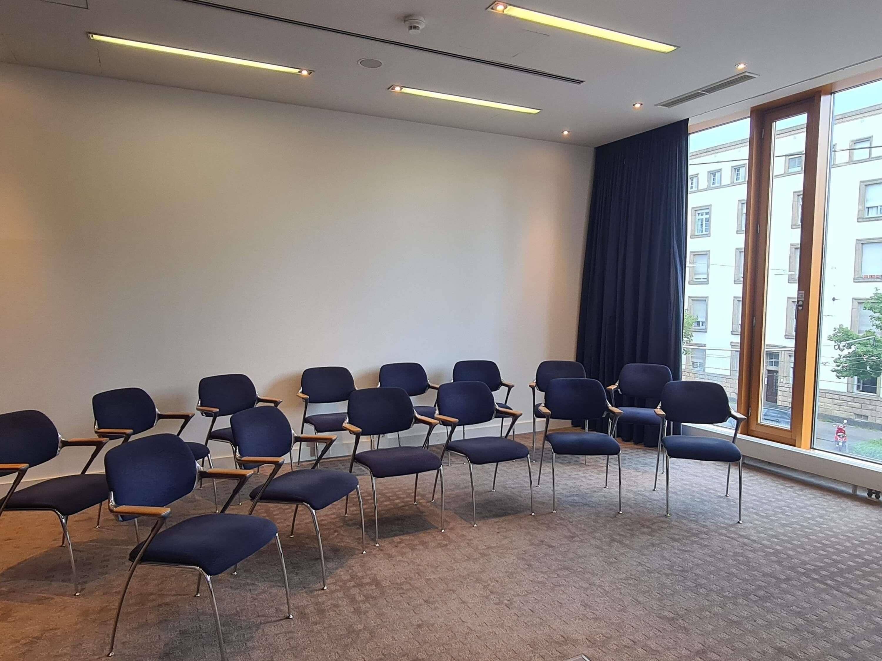 A conference room with rows of chairs, a large window with a view of buildings, and ceiling lights.