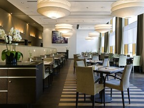 Un comedor moderno de hotel con varias mesas y sillas, iluminación colgante y un mostrador de recepción.
