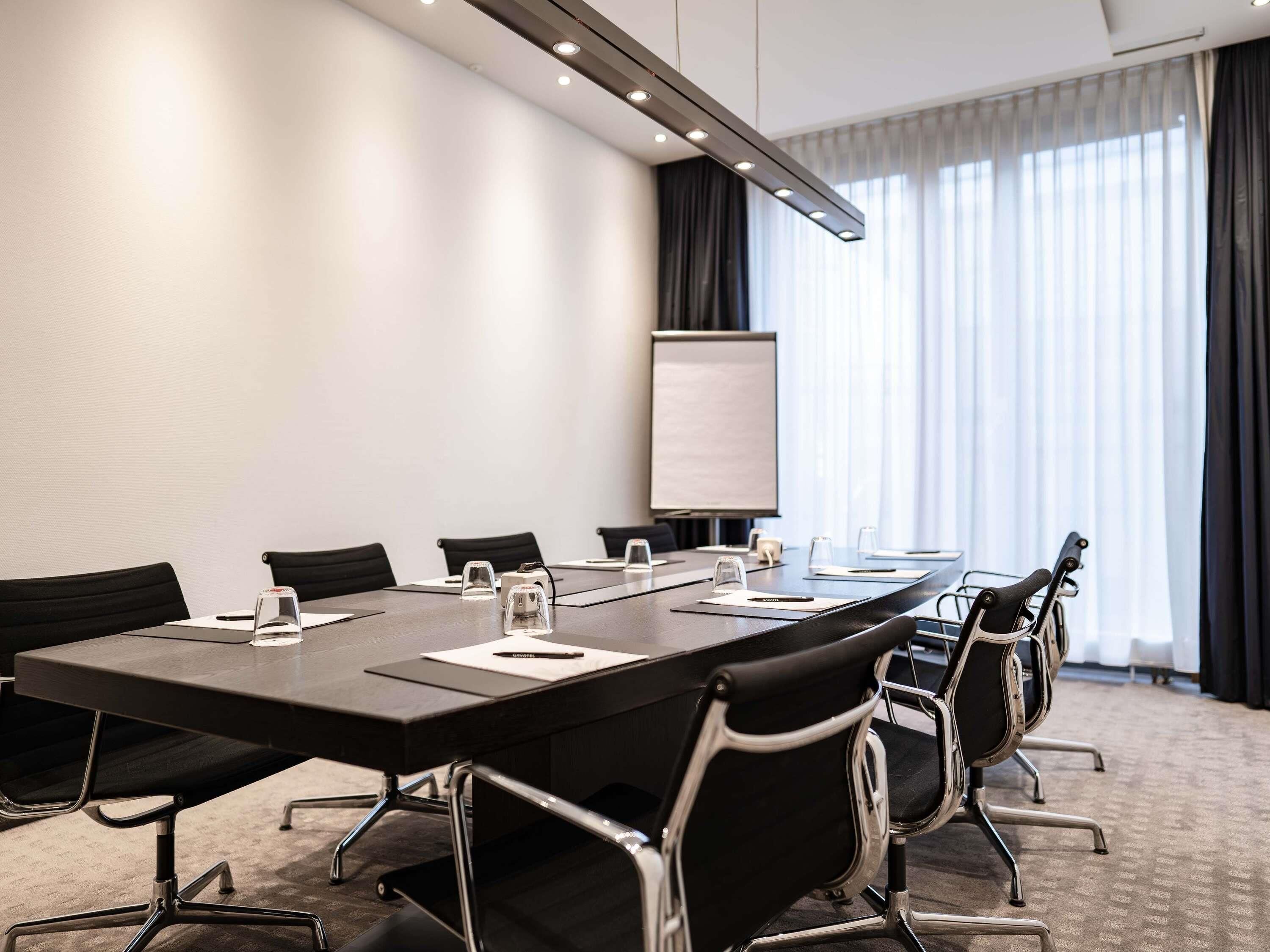 A conference room with a long table, black chairs, a whiteboard, and a window with curtains.