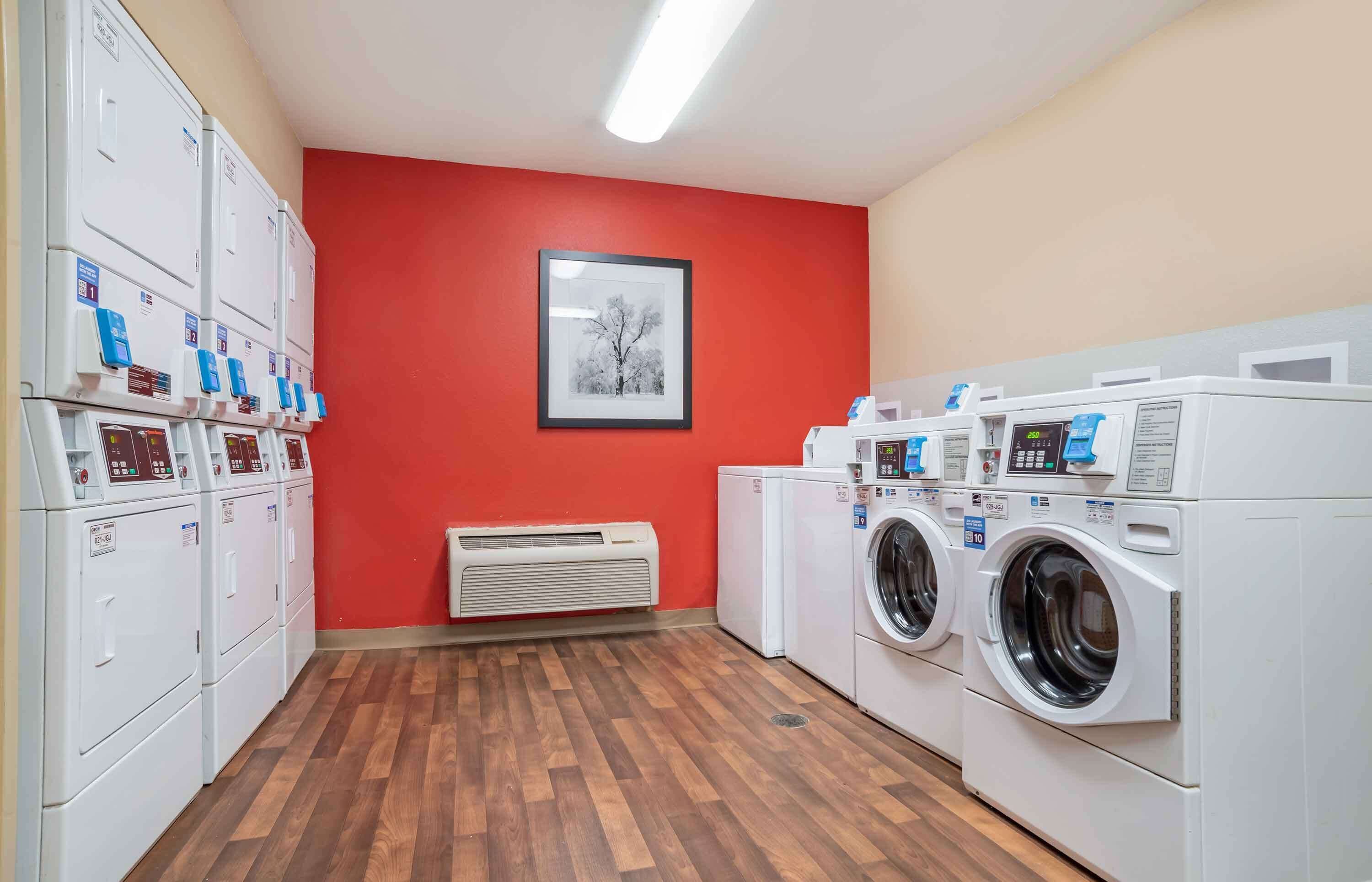 A laundry room with multiple washing machines and dryers, a framed picture on the wall, and a red wall.