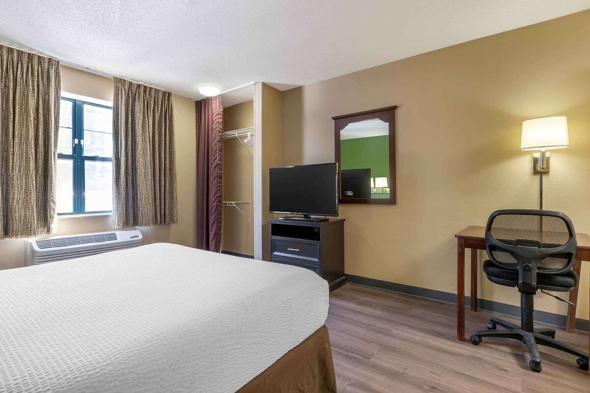 Ett hotellrum med en säng, ett skrivbord med en stol, en tv, en spegel och ett fönster med gardiner.