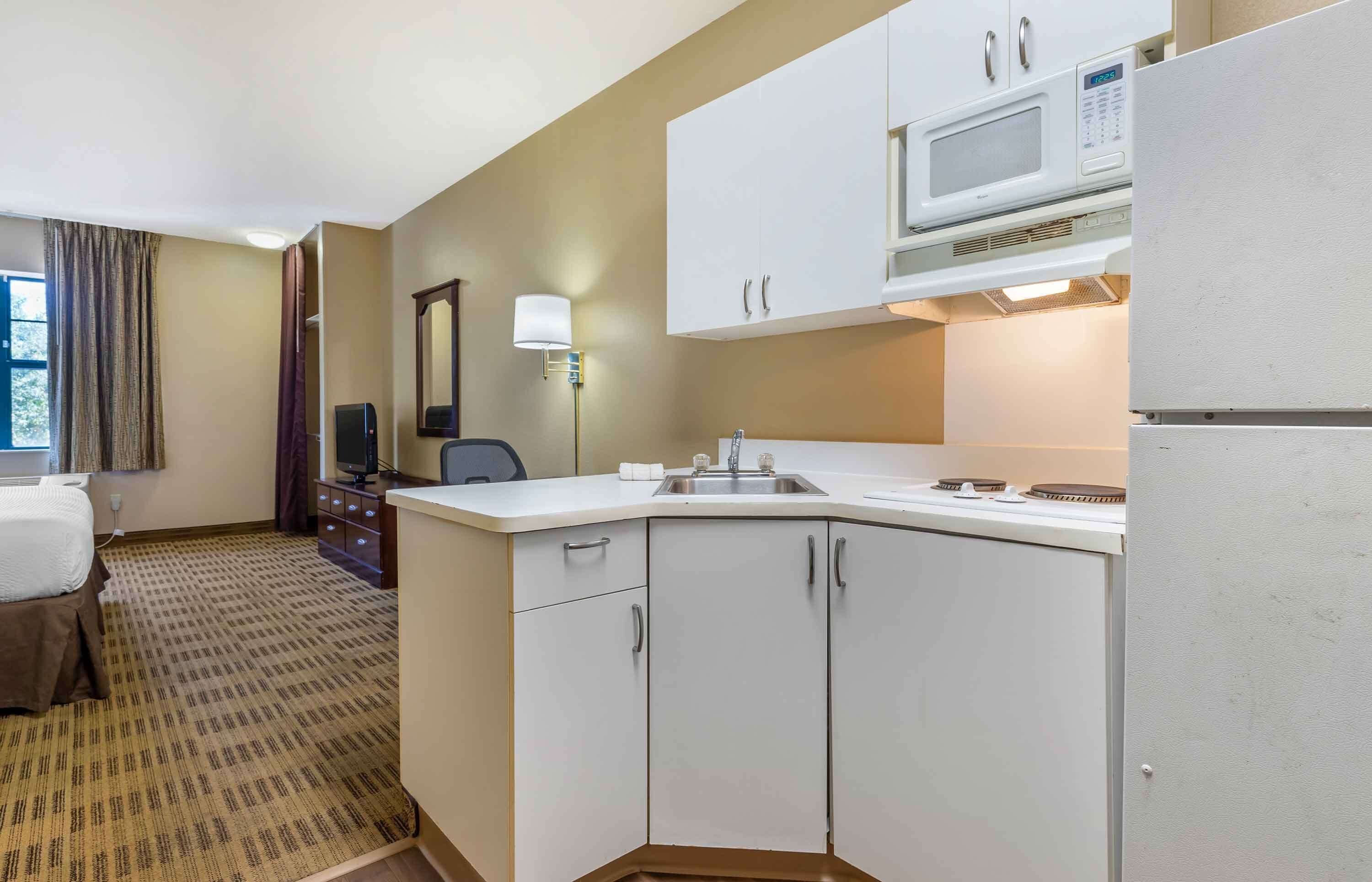 A compact hotel room with a kitchenette, a bed, a desk, and a window with curtains.
