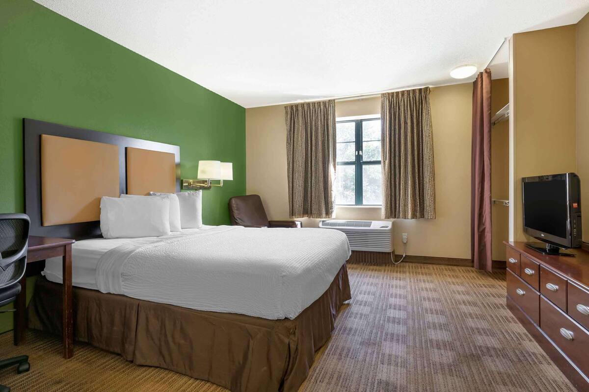 Ett hotellrum med en stor säng, ett skrivbord med en stol, en tv, ett fönster med gardiner och en luftkonditioneringsanläggning.