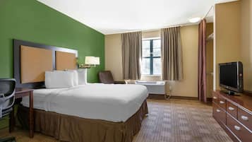 A hotel room with a large bed, a desk with a chair, a television, a window with curtains, and an air conditioning unit.
