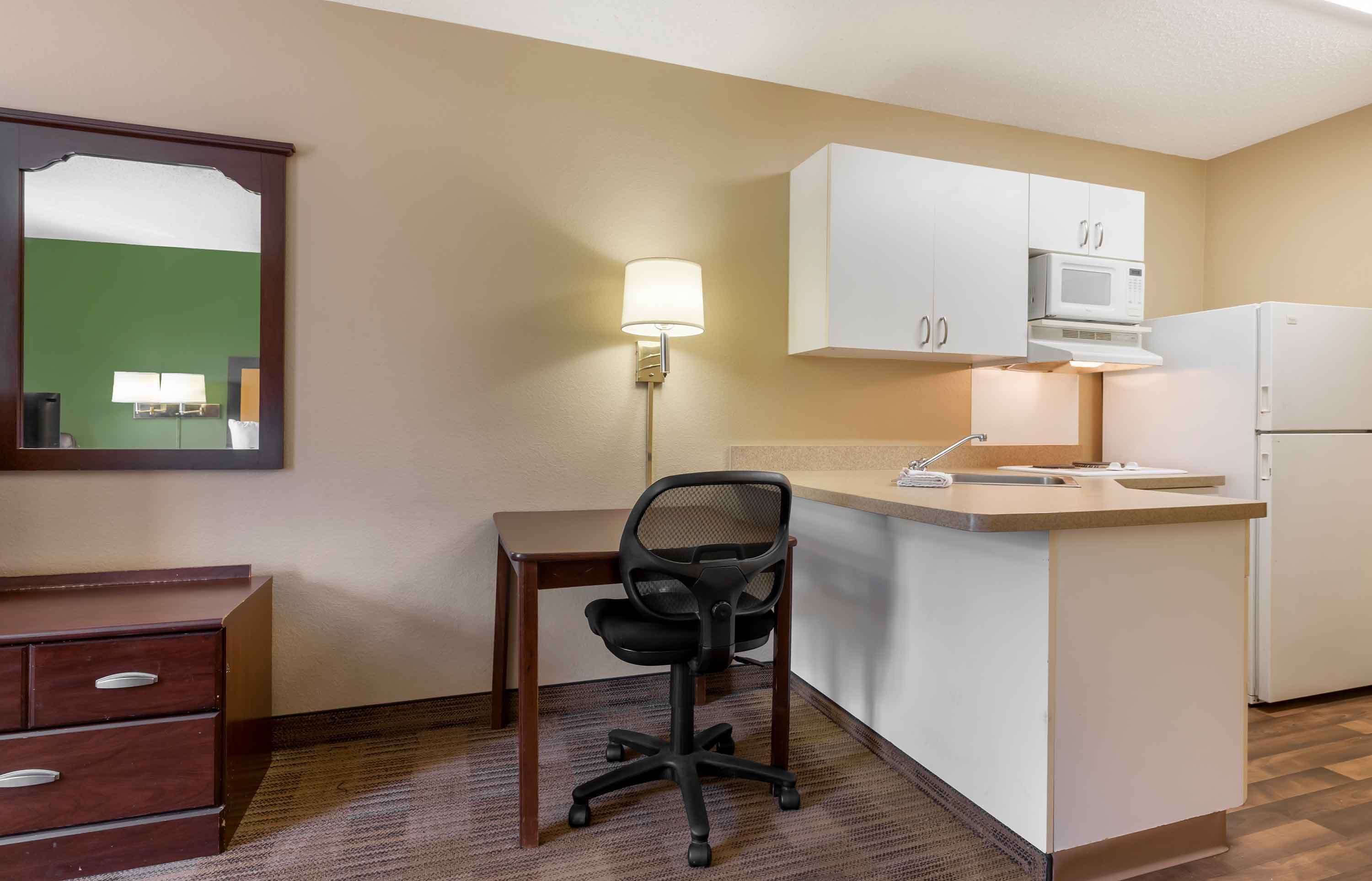 A compact hotel room with a kitchenette, a desk with a chair, a mirror, and a lamp.