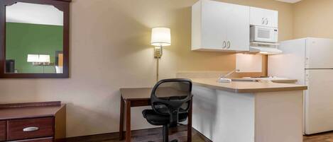 A compact hotel room with a kitchenette, a desk with a chair, a mirror, and a lamp.