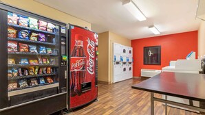 Une machine distributrice de collations et un réfrigérateur à boissons, notamment des Coca-Cola, une table et un espace pour faire la lessive, le tout dans une pièce au sol en bois et aux murs rouges.