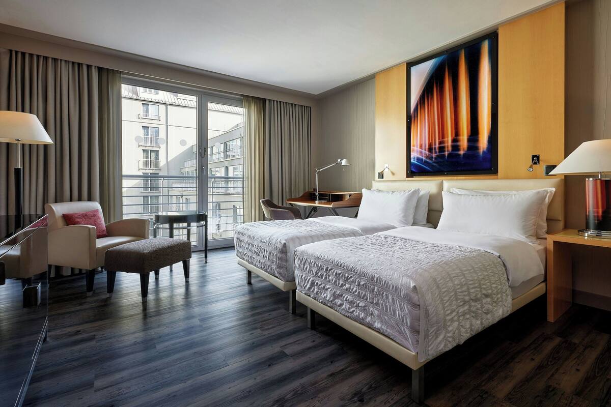 Hotellihuone, jossa on suuri sänky, työpöytä, tuoli ja ikkunasta avautuva näkymä ympäröiviin rakennuksiin.