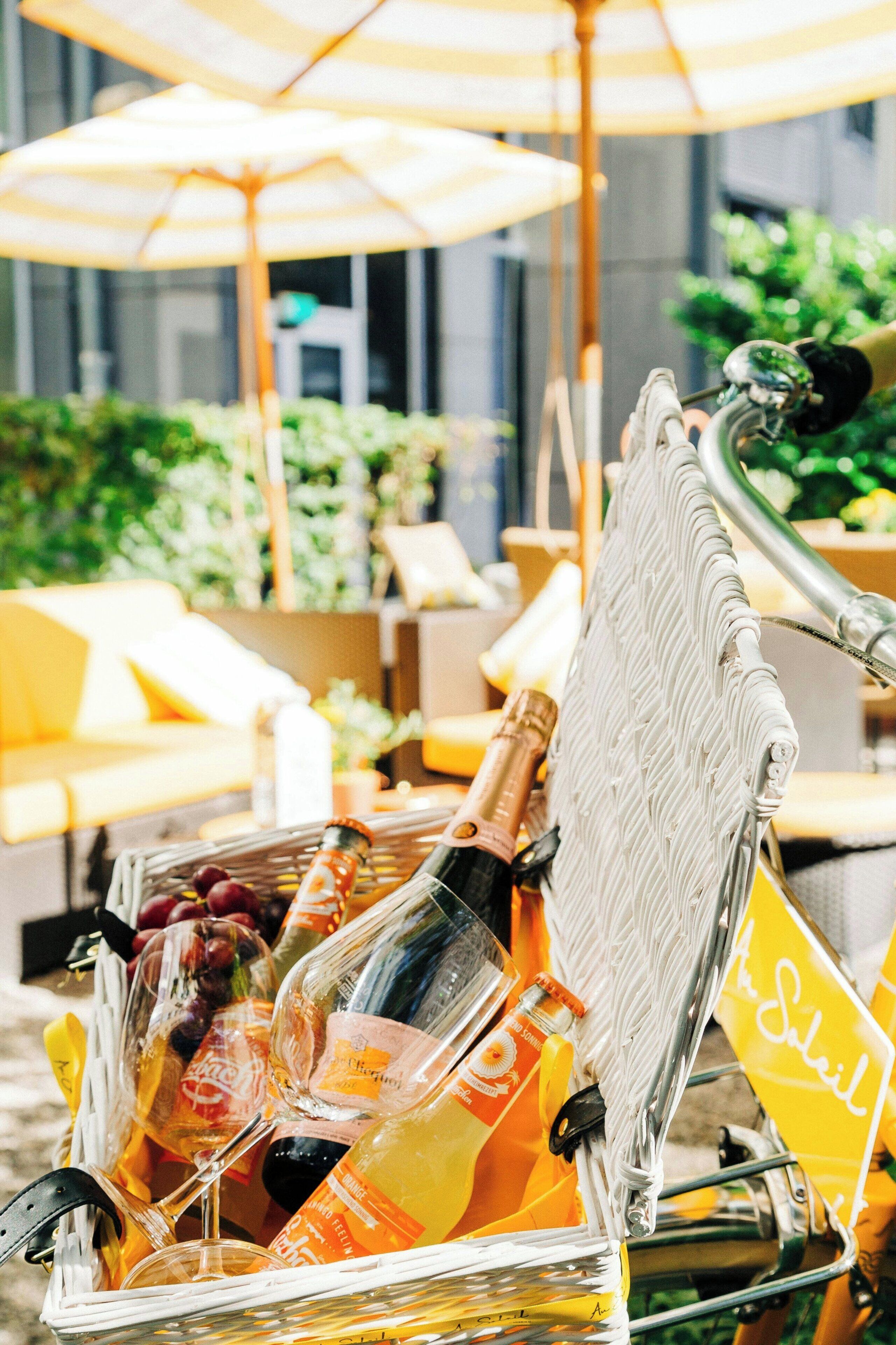 A bicycle with a basket containing bottles and fruits, set outdoors with umbrellas and greenery in the background.
