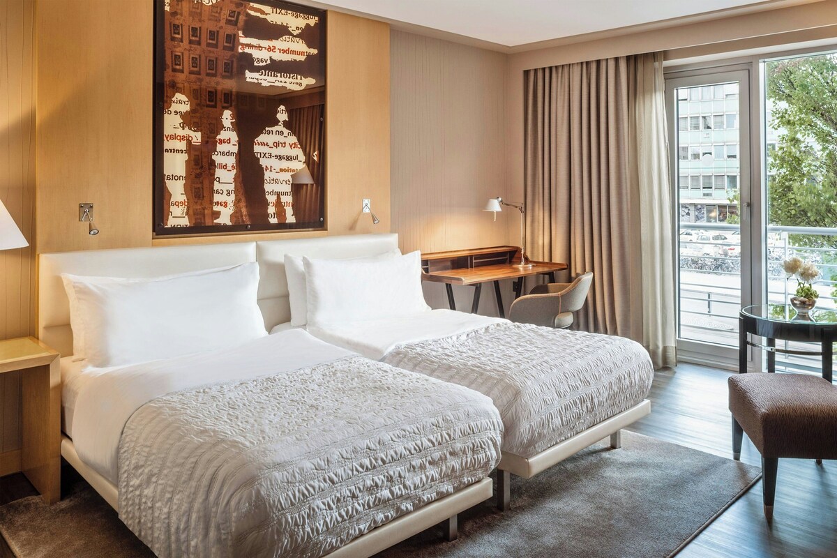 A hotel room with two beds, a desk, a chair, and a large window with a view of buildings and trees.