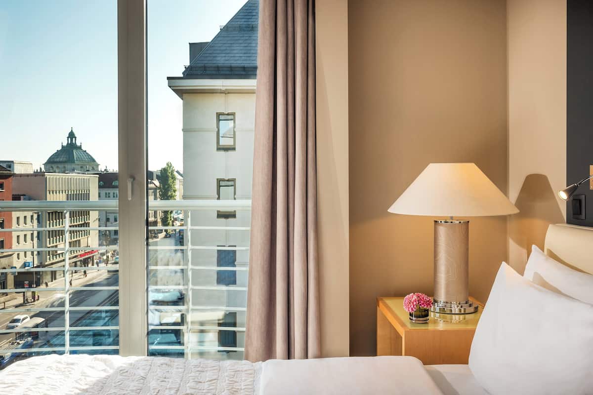 Hotellihuone, jossa on suuri ikkuna, sänky, yöpöytä lampun kanssa ja näkymä kaupungin rakennuksiin.