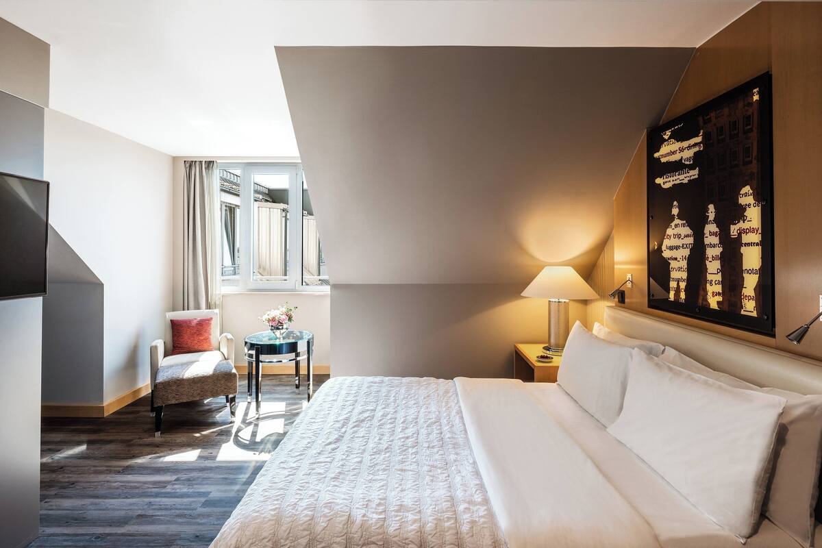 Hotellihuone, jossa on suuri sänky, tuoli, pieni pöytä, lamppu ja seinälle ripustettu taideteos.
