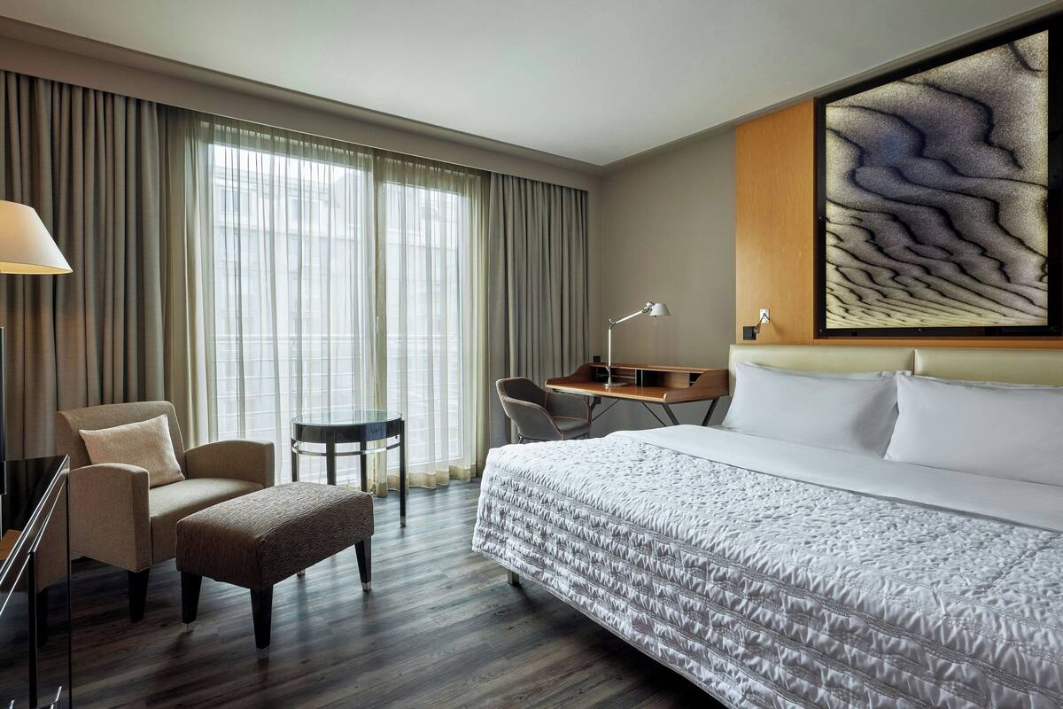 Hotellihuone, jossa on suuri sänky, työpöytä, tuoli ja ikkunasta avautuva näkymä kaupunkiin.