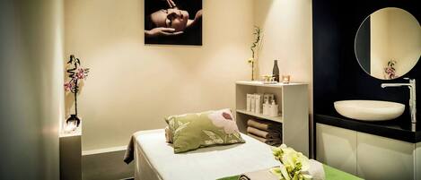A modern spa room with a bed, a large mirror, a sink, and a shelf with toiletries.