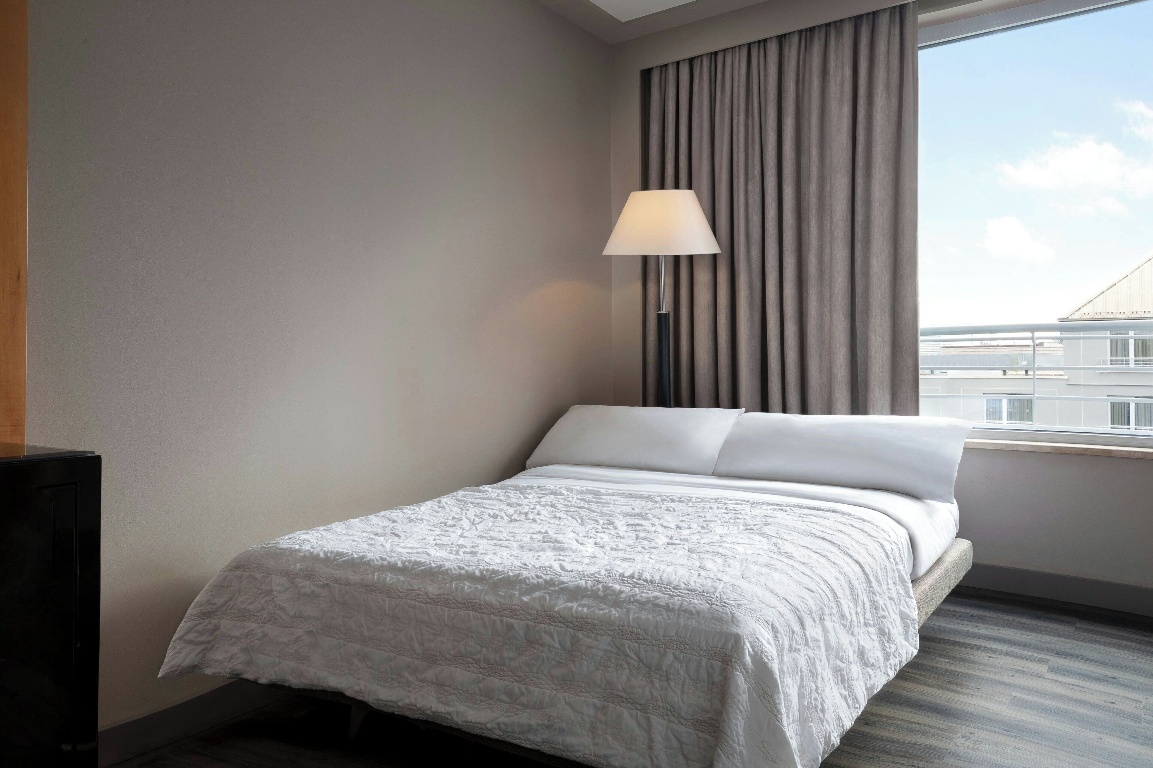 A hotel room with a bed, a lamp, and a view of buildings through the window.