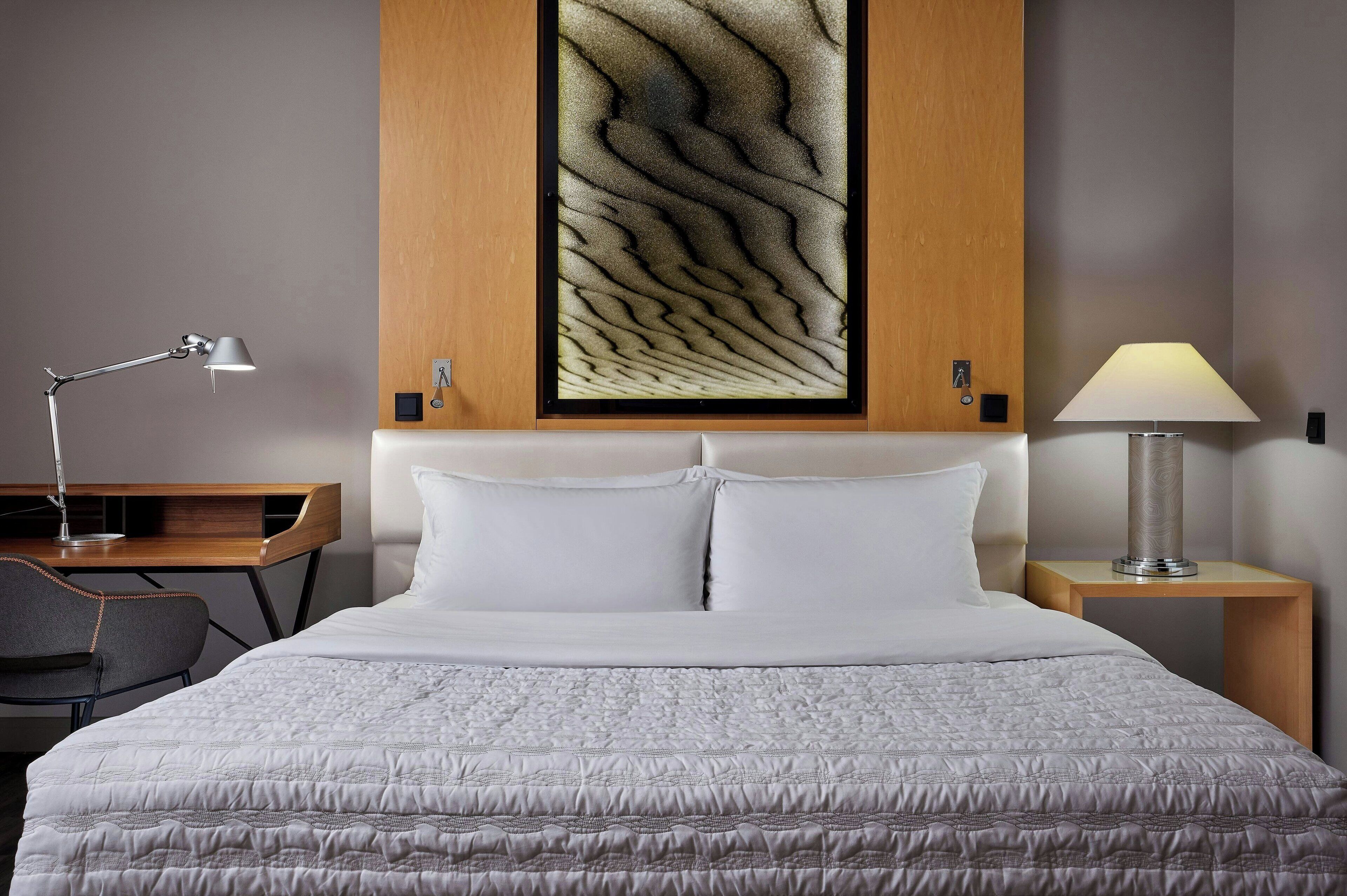 A neatly made bed with white bedding, a desk with a lamp, and a wall-mounted artwork.