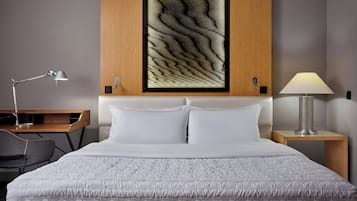 A neatly made bed with white bedding, a desk with a lamp, and a wall-mounted artwork.
