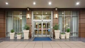 Entrada de hotel com porta de vidro, plantas em vasos e tapete azul.