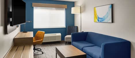 A modern hotel room with a blue sofa, a wooden desk, a television, a lamp, and a radiator.