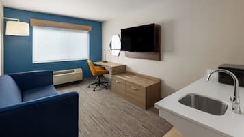 A hotel room with a blue sofa, a wooden desk, a TV, and a kitchen area with a sink.