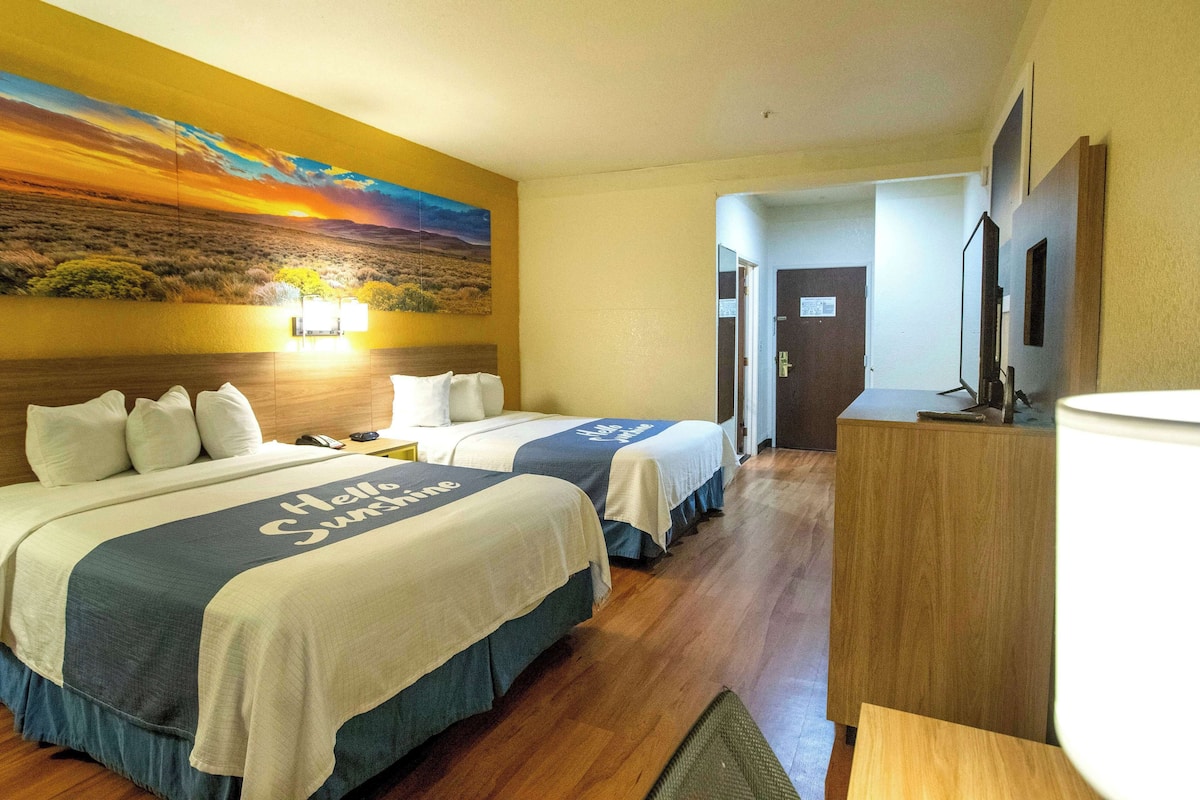 Ein Hotelzimmer mit zwei Betten, einem Sonnenuntergangsbild, einem Schreibtisch und einem Spiegel.