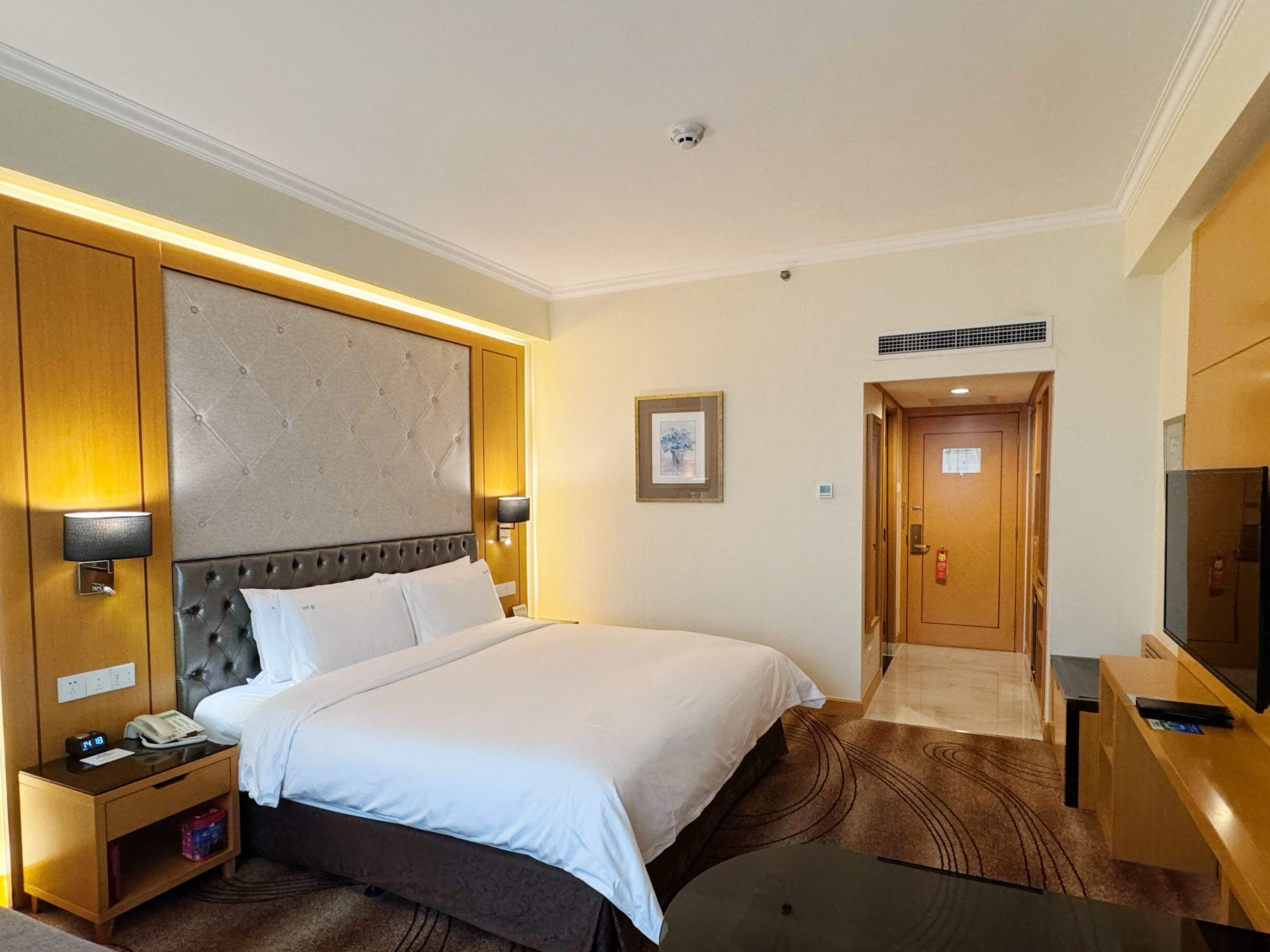 Una habitación de hotel con una cama grande, una mesita de noche con teléfono y reloj, un escritorio con televisor, y una puerta que da a otra habitación.