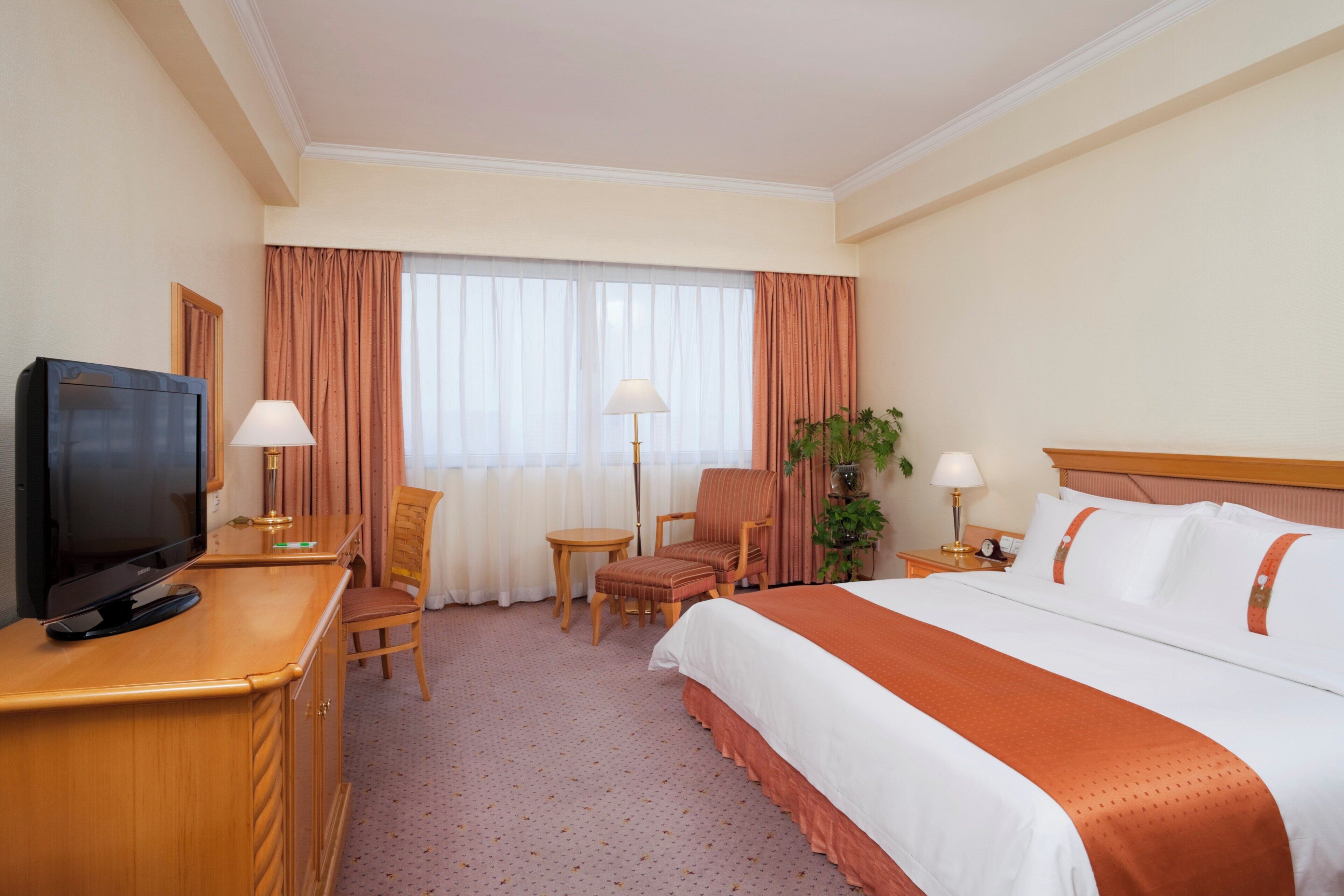 Habitación de hotel con cama, escritorio con televisor, una silla, una mesita y ventana con cortinas.