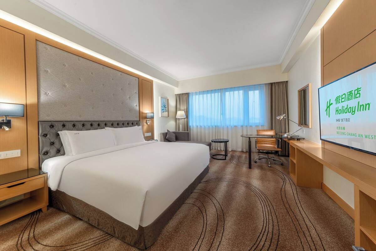 A hotel room with a large bed, a desk, a chair, a TV, and a view of the city.