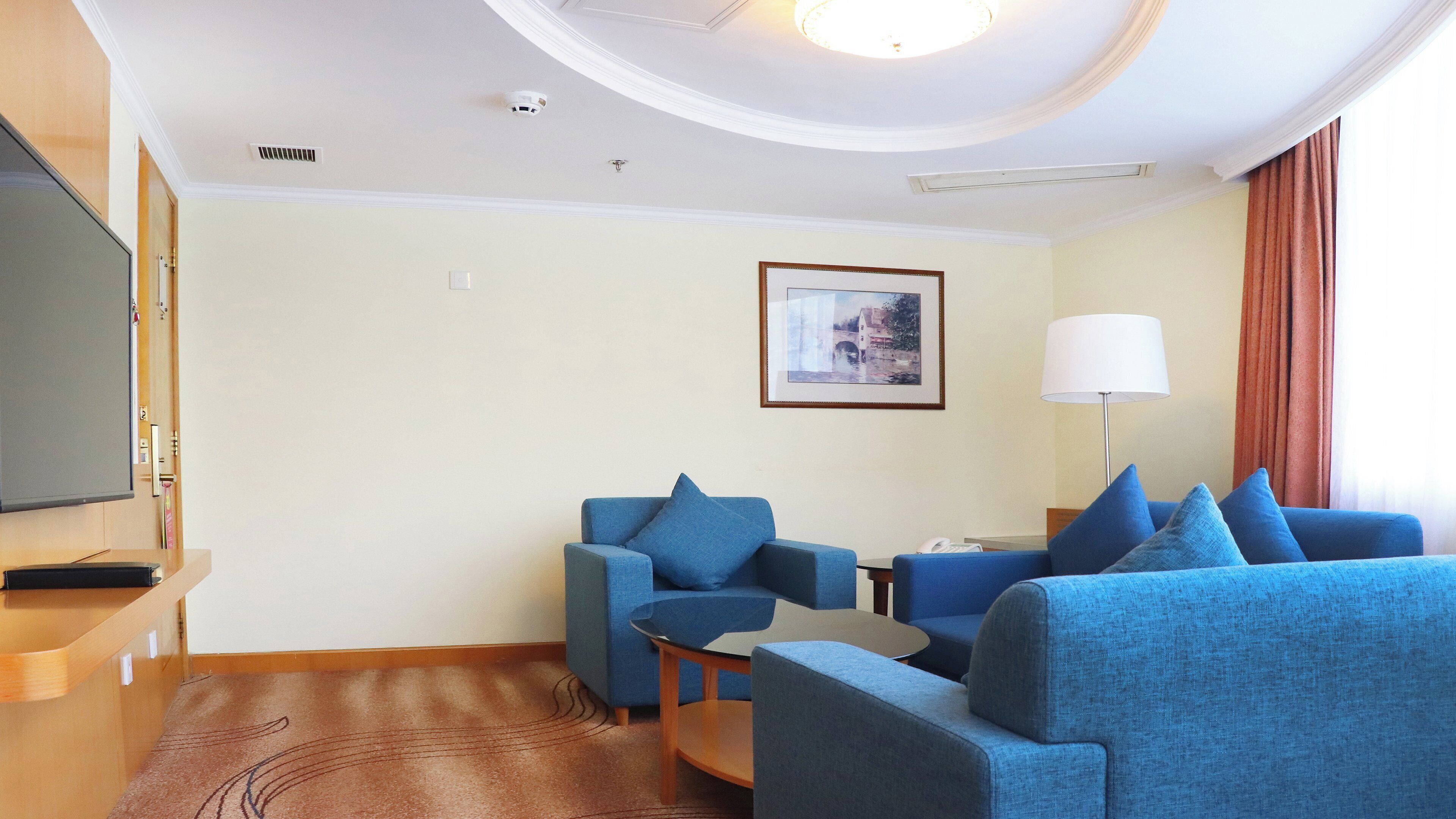 A hotel room with two blue sofas, a flat-screen TV, a framed picture on the wall, and a lamp.