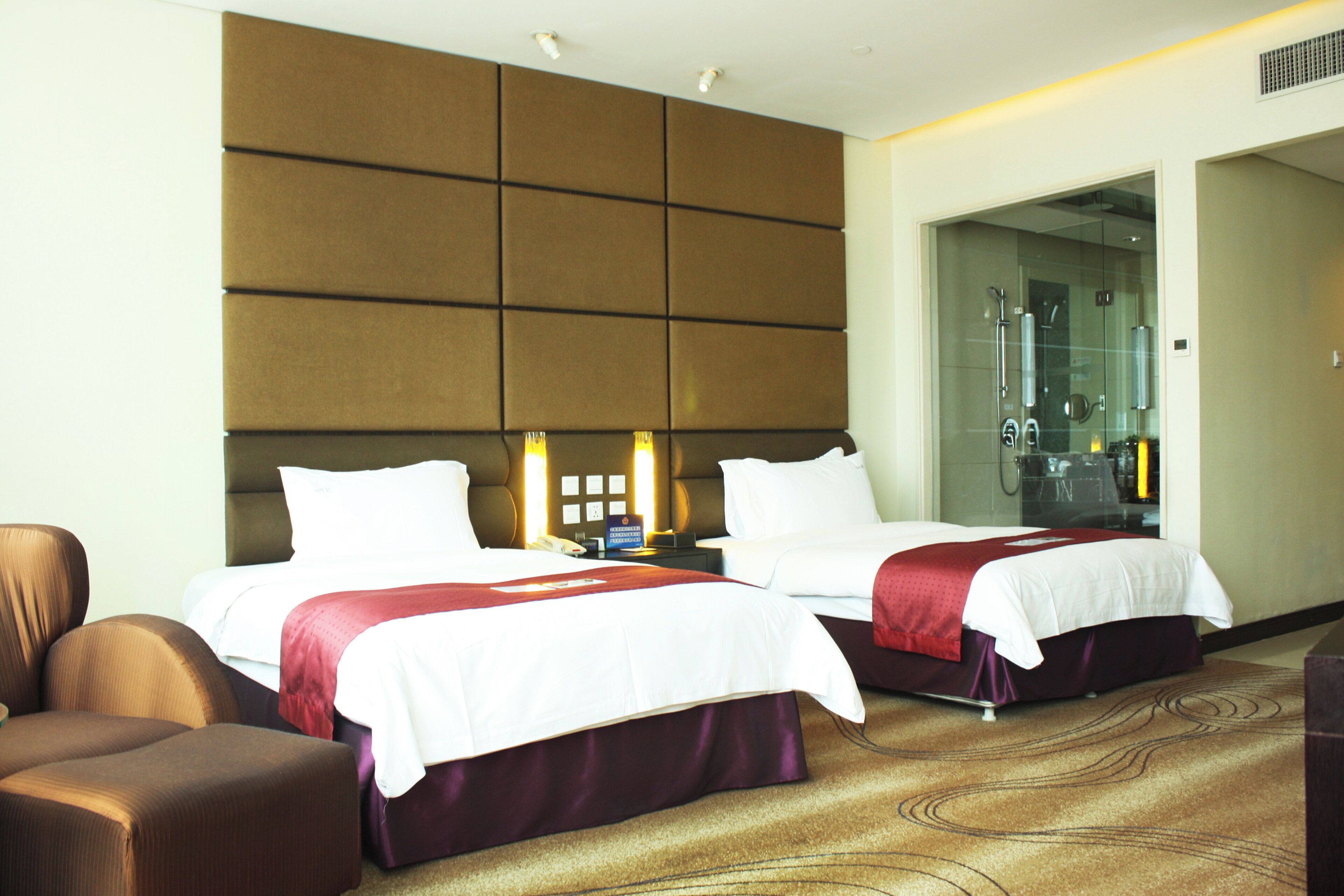 A hotel room with two beds, a sofa, a bathroom with a glass door, and a wall-mounted control panel.