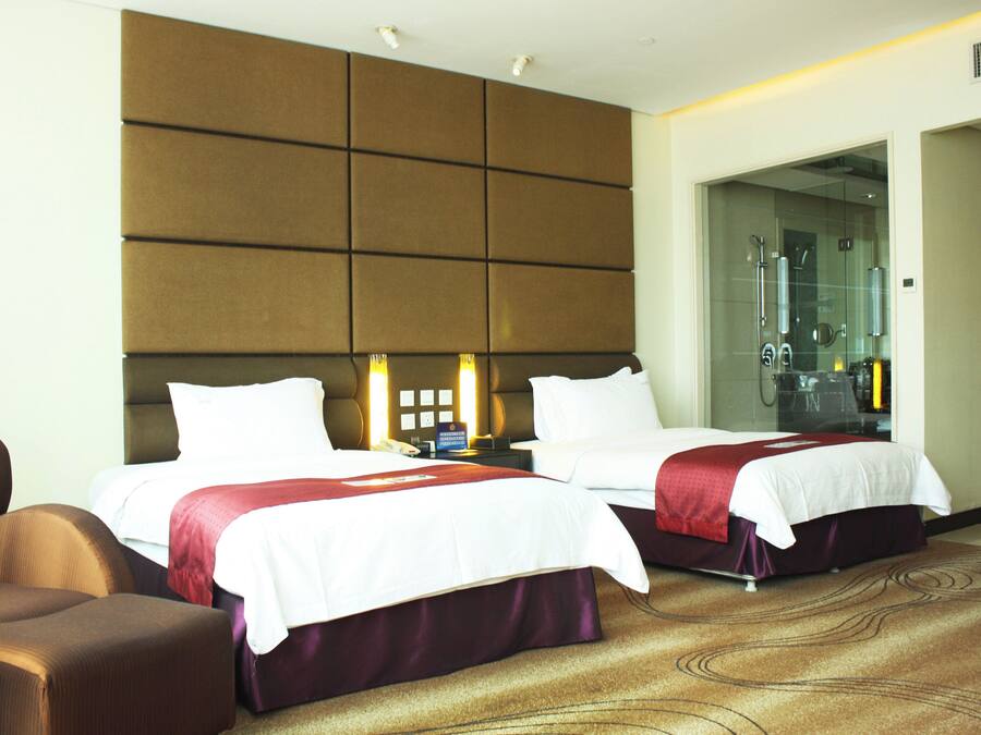 A hotel room with two beds, a sofa, a bathroom with a glass door, and a wall-mounted control panel.