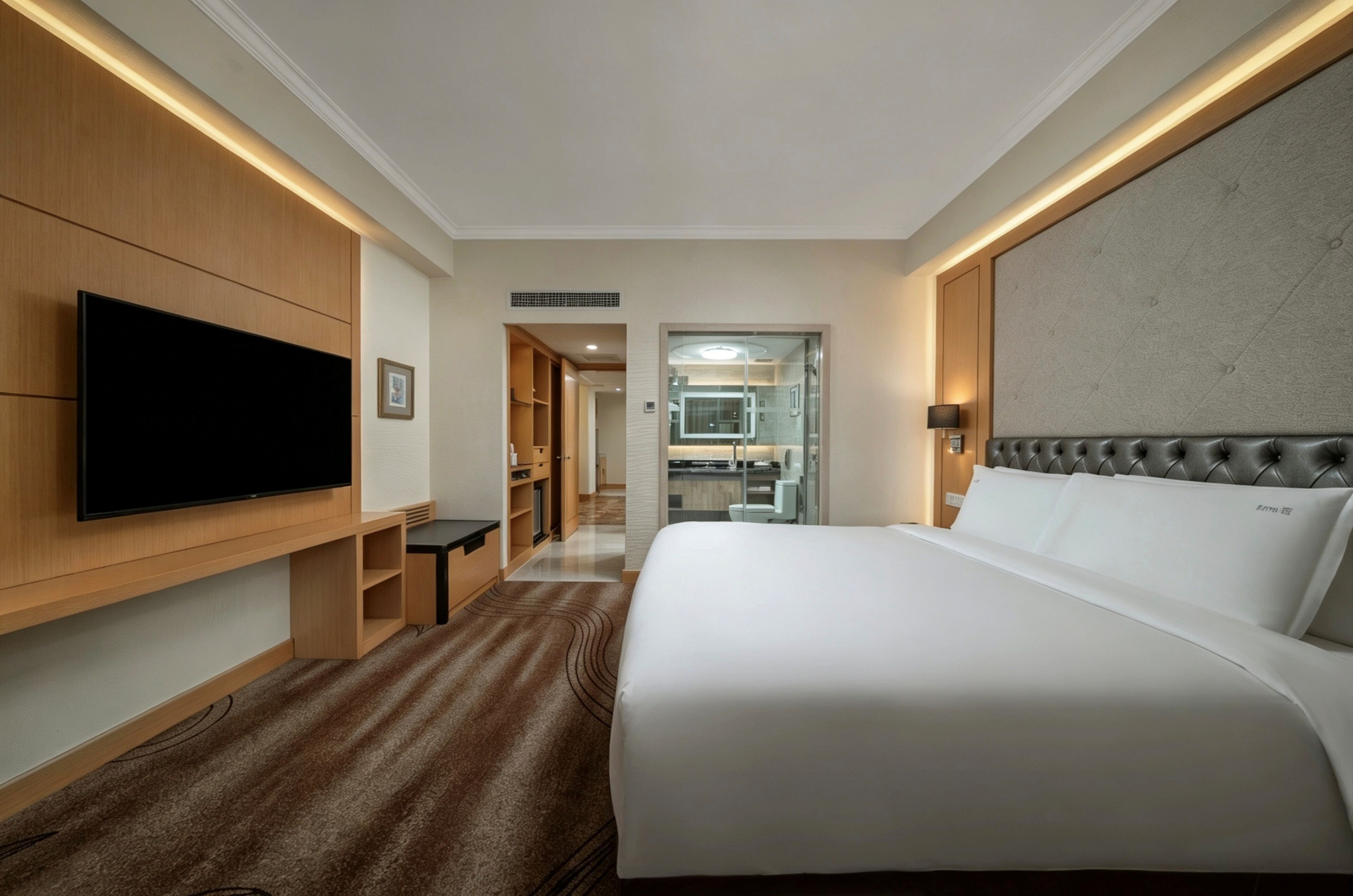 A hotel room with a large bed, a flat-screen TV mounted on the wall, a wooden wardrobe, and a bathroom visible through an open door.