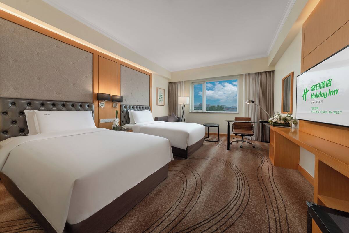 A hotel room with two beds, a desk, a chair, a TV, and a window with a city view.