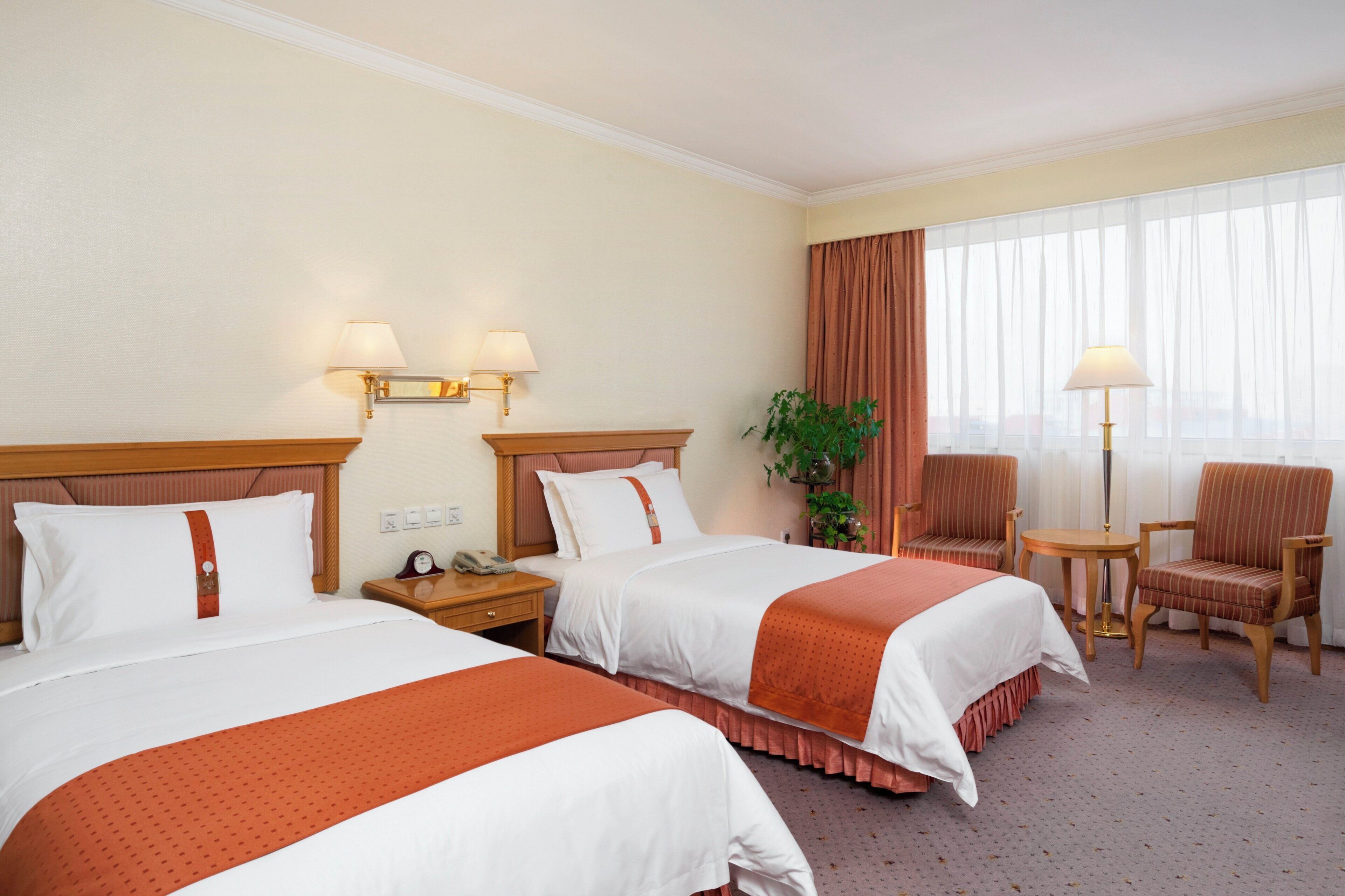 A hotel room with two beds, a nightstand, a chair, a table, and a large window with curtains.