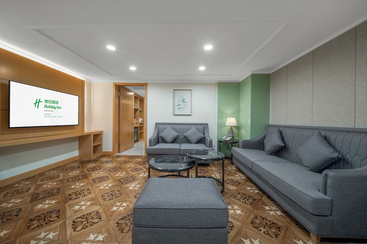 A hotel room with a grey sofa, a grey ottoman, a small table, a TV mounted on the wall, and patterned flooring.